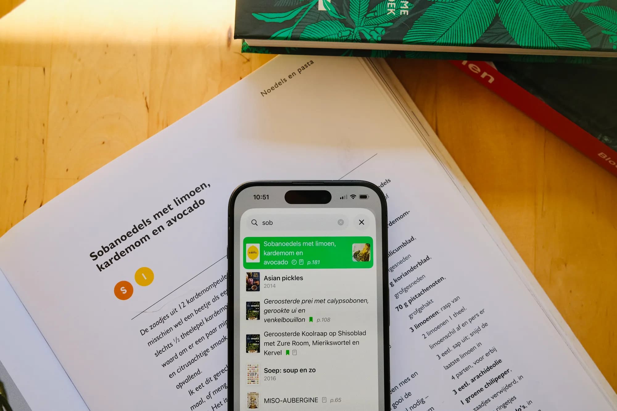 Een iPhone die in CookCook zoekt naar sobanoedels, liggend op het opengeslagen kookboek waar het recept in staat